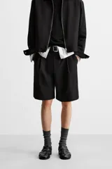 Bermuda de corte relajado color negro, con pliegues frontales, cierre de cremallera y botón, bolsillos laterales y bolsillos traseros.