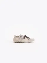 Zapatillas de cuero vacuno en color beige, con recortes de gamuza en la puntera y talón, doble cordón en tonos gris y marrón, hebilla lateral y dije colgante en la cordonera. Suela plana de goma.