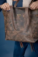 Bolso tipo shopper de cuero color suela con textura grabada, detalle de costuras laterales trenzadas y placa metálica con logo de la marca. Cuenta con doble asa larga para colgar al hombro, cierre principal y múltiples bolsillos internos y externos.