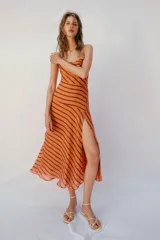 Vestido midi de color naranja con rayas diagonales marrones, escote fluido y tirantes finos cruzados en la espalda con abalorios. Presenta corte evasé con abertura frontal y forro interior.