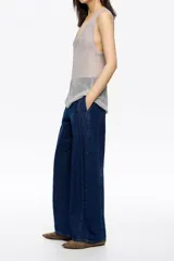 Pantalón de denim azul oscuro, corte barrel (tobillero con volumen en la cadera y estrechamiento hacia el tobillo). Presenta tiro medio, cierre frontal con corchete oculto y cremallera, y tiras ajustables con hebilla en la espalda.