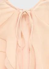 Blusa de viscosa color beige, con diseño de volantes frontales, cuello escotado con cierre de lazo y mangas largas acampanadas con volantes en los puños.