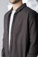 Camisa verde militar de corte recto, con cuello clásico, cierre frontal con botones a presión, mangas largas con puños abotonados, dos bolsillos de parche en el pecho y bajo redondeado.