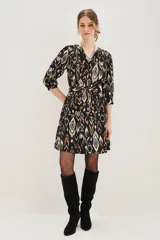 Vestido corto de corte recto con estampado ikat en tonos negro, blanco y ocre. Presenta escote en V con cierre de botones, mangas tres cuartos con puños elásticos y un cinturón de la misma tela para ajustar la cintura.