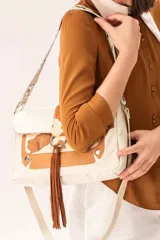 Cartera de cuero vacuno en tono arena con detalles en cuero camel y apliques de pelo vacuno estampado. Presenta un diseño con solapa frontal, fleco central de gamuza, herrajes metálicos y cierre principal. Incluye una correa corta con tachas y una correa larga extensible y desmontable.