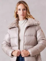 Campera puffer larga de acabado satinado en tono beige. Presenta un diseño capitoneado horizontal, cierre frontal bidireccional y un amplio cuello envolvente revestido en piel sintética. Incluye bolsillos laterales funcionales con cierre y logo metálico en la manga.