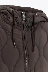 Chaqueta acolchada de color marrón oscuro con diseño de costuras onduladas, cuello subido, capucha ajustable con cordones, cierre frontal con cremallera y cintura ajustable con cordones. Cuenta con bolsillos delanteros de vivo y tejido repelente al agua.