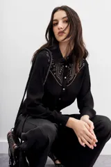 Camisa de estilo texano en color negro, con mangas largas, cuello camisero y cierre frontal con botones metálicos. Presenta un diseño con tachas termo fijadas en la zona del canesú delantero.
