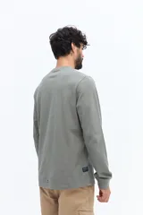 Remera de manga larga color blanco roto o gris muy claro, con escote redondo y un pequeño logo circular estampado en el pecho.