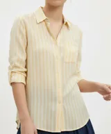 Camisa de lino y viscosa color amarillo pastel con rayas verticales blancas. Presenta cuello clásico, cierre frontal de botones y bolsillo de parche en el pecho. Las mangas son largas y se pueden remangar con presilla.