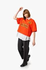 Remera naranja con cuello polo blanco, estampado frontal con la palabra "GOAL" en amarillo y negro, y logos bordados.