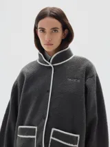 Campera de sherpa color gris oscuro con detalles de vivos en color blanco. Presenta cuello alto, cierre frontal con broches a presión, mangas largas y bolsillos frontales con borde a contratono. Incluye el logo de la marca bordado en el pecho.
