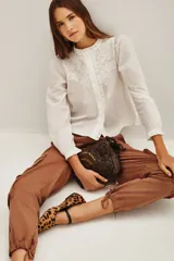 Camisa blanca de algodón con cuello alto, cierre frontal de botones ocultos y detalles de bordado floral en el pecho. Presenta mangas largas con puños elásticos y volados decorativos en la parte frontal.