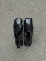 Bota corta de punta cuadrada con acabado efecto cocodrilo y paneles laterales elásticos. Presenta una costura central visible y una suela plana con detalle metálico en la puntera.
