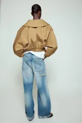 Chaqueta corta de Zara confeccionada en mezcla de algodón, con cuello solapa, manga larga con hombro caído y puños elásticos. Presenta un diseño cruzado con cierre de hebilla metálica lateral y forro interior a tono.
