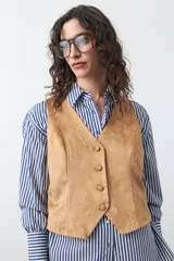 Chaleco de gamuza color marrón, con escote en V y cierre frontal con botones forrados. Tiene un largo a la cintura y silueta definida.