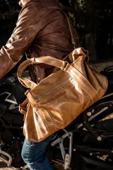 Bolso de cuero hecho a mano en color tostado, con diseño amplio y funcional. Presenta cierre superior, bolsillo frontal con cremallera, asas de mano reforzadas y una correa larga ajustable y desmontable.