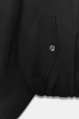Chaqueta bomber negra con cuello redondo y pañuelo desmontable anudado al frente. Presenta cierre frontal con cremallera, bolsillos laterales con solapa y botones a presión, y terminaciones elásticas abullonadas en puños y ruedo.
