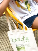 Bolso tote color beige claro con asas de tela, presentando un estampado gráfico de un vaso de jugo de matcha con capas de colores verde, blanco y naranja, y texto que dice "matcha juice".