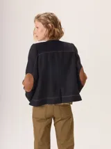 Chaqueta de paño color marrón oscuro, de corte clásico y cuello redondo, con cierre frontal dorado. Presenta bolsillos rectangulares con ribetes en color rojizo y parches de pana a rayas en los hombros.