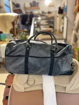 Bolso de viaje tipo duffel de cuero negro con detalles de refuerzo en gamuza y doble asa de mano.