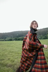 Capa oversize de corte amplio con estampado de cuadros en tonos rojos, negros y marrones. Presenta cuello alto, cierre frontal con botones, bolsillos delanteros y mangas ajustables mediante trabillas con botones.