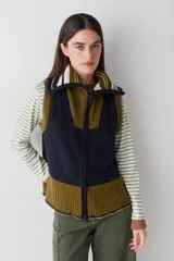 Chaleco de lana con cierre frontal completo y cuello alto. Presenta un diseño color block que combina paneles en azul marino con tejido de ochos y paneles en verde oliva con tejido acanalado. Incluye detalles de rayas en el cuello y un corte asimétrico más largo en la parte posterior con aberturas laterales.