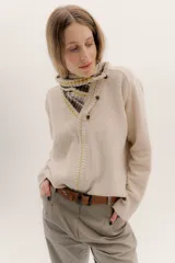 Polera tejida en lambswool color beige, con cuello alto y diseño de intarsia en tonos marrones y grises. Presenta botones forrados en el cuello para modular la apertura.