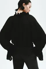Blusa oversized color negro en tejido liviano de mezcla de lino y viscosa, con cuello elevado plisado y abertura en V con tiras finas. Mangas raglán extralargas con detalles bordados tono sobre tono y puños acampanados con pliegues.
