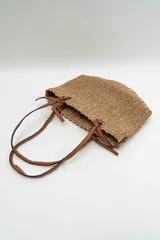 Bolso de mano tipo tote confeccionado en tejido texturizado efecto rafia en color beige, con asas dobles finas de cuero sintético marrón y detalles de nudos en los extremos.