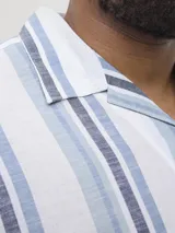 Camisa de manga corta con cuello resort y ajuste relajado. Presenta un diseño de rayas verticales en tonos azules y blancos, con cierre frontal de botones. Confeccionada en una mezcla de algodón y lino, ofrece una textura ligera y fresca.