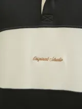 Buzo de manga larga con cuello tipo polo y fit relajado, en color negro con una franja horizontal beige en el pecho. Presenta puños acanalados y un bordado en el centro de la franja beige.