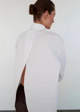 Camisa blanca de corte oversize y diseño largo, confeccionada en algodón. Presenta cuello camisero, cierre frontal con botones, mangas largas con puños abotonados y un distintivo diseño de espalda cruzada.