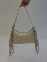 Cartera de hombro color beige con flecos laterales decorativos y correa regulable con argollas metálicas. Cuenta con cierre superior y bolsillo interno.