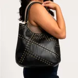 Cartera al hombro Korium modelo Aster, de color negro, con diseño estructurado y detalles de tachas metálicas doradas y plateadas distribuidas en líneas geométricas. Cuenta con doble asa de hombro.