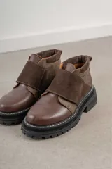 Borcego de caña baja en combinación de cuero liso y gamuza color marrón, con cierre de velcro ajustable en el empeine y suela track dentada negra.