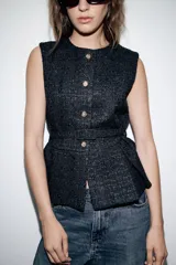 Chaleco de tejido tipo tweed en color negro, con cuello redondo y cierre frontal de botones dorados. Presenta un diseño entallado con cinturón integrado del mismo tejido y bajo con pliegues.