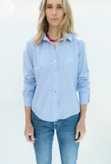 Camisa de popelina a rayas celestes y blancas, de estilo clásico, con cuello camisero, manga larga con puños ajustados, bolsillo frontal aplicado y botones de nácar.