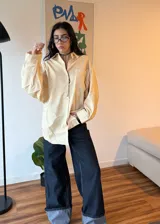 Camisa oversize de corte amplio con estampado de rayas verticales finas en tonos amarillo y azul. Presenta cuello clásico, cierre frontal con botones, mangas largas con puños abotonados y un bolsillo en el pecho.
