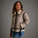 Campera estilo aviador confeccionada en gamulán color marrón claro, forrada internamente con corderito sintético. Presenta cuello, puños y ruedo con detalles de corderito a la vista, cierre frontal metálico y cinturón ajustable en la cintura con hebilla. Incluye detalles de tiras en cuero sintético negro en costuras y cierres.