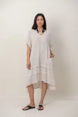 Vestido midi de lino color beige, de corte holgado y silueta acampanada. Cuenta con cuello redondo, mangas cortas amplias, bolsillos laterales y detalles de recortes horizontales en la falda con terminación asimétrica más larga en la parte trasera.