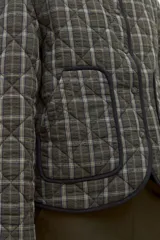 Campera capitoneada de corte recto con diseño a cuadros en tonos verde oliva y gris. Presenta cuello camisero de pana en color negro, cierre frontal con botones a presión y bolsillos laterales amplios con ribete negro.