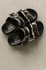 Sandalias de cuero de doble tira, con estampa animal print tipo vaca en tonos blanco, negro y marrón, adornadas con tachas metálicas en la tira superior y una hebilla grande en la tira inferior.