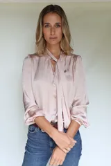 Camisa de seda color rosa, con cuello a la base y lazos frontales integrados. Presenta mangas con leve frunce en los hombros, puños elásticos y botones de nácar.