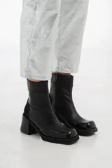 Bota corta de cuero negro con diseño bicolor que combina cuero liso y cuero charol en la puntera. Cuenta con taco cuadrado de altura media y cierre lateral interno.