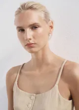 Chaleco tipo crop top de color beige, con tirantes finos, escote recto y cierre frontal de botonadura con botones revestidos. Cuenta con dos bolsillos de ribete delanteros y bajo con diseño asimétrico.