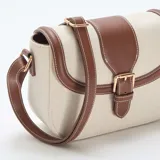 Bolso bandolera de diseño estructurado, confeccionado en lona color beige con detalles de cuero sintético marrón en los bordes y la solapa frontal. Cuenta con una hebilla decorativa metálica, costuras a la vista en color blanco y correa ajustable.