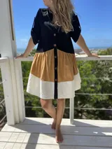 Vestido de lino en tres colores: negro en la parte superior, ocre en el medio y blanco en la parte inferior. Tiene botones en el frente y mangas cortas con detalle de lazo.