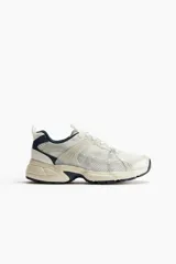 Zapatillas deportivas blancas con detalles en gris y azul marino. Capellada de malla con refuerzos sintéticos, cuello y lengüeta acolchados, cierre con cordones y suela gruesa con diseño.