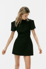 Vestido mini negro con cuello redondo, manga corta con pliegues y costuras marcadas. Cuenta con cierre posterior con cremallera oculta.